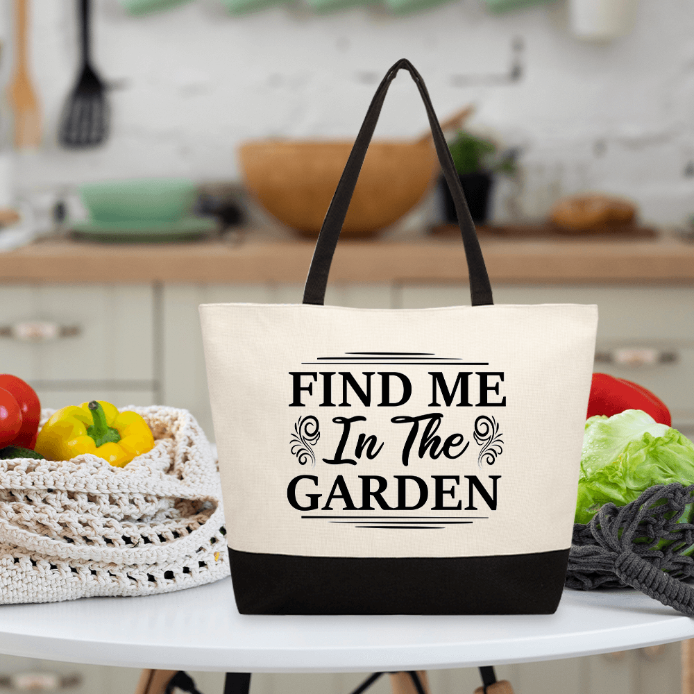 A canvas tote bag with black handles and the text "FIND ME In The GARDEN" printed on the front, placed on a white table with vegetables and kitchen utensils in the background.
