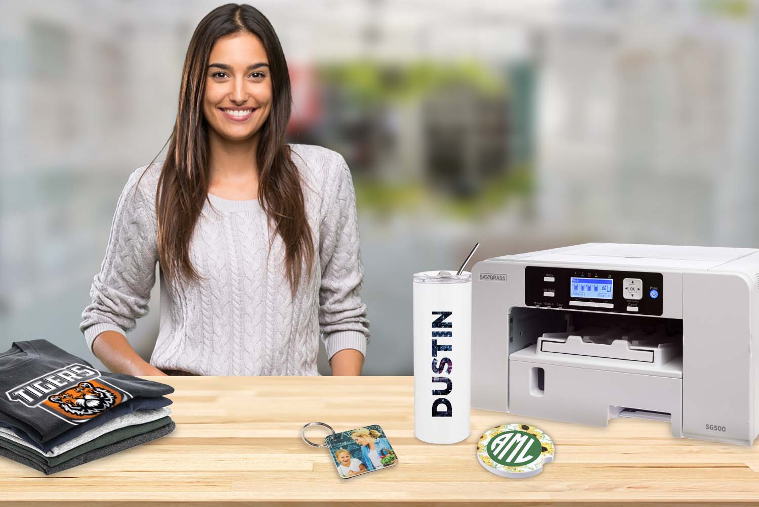 Woman behind a table holding a printer, shirts, a keychain, a coaster, and a tumbler that are all personalized.